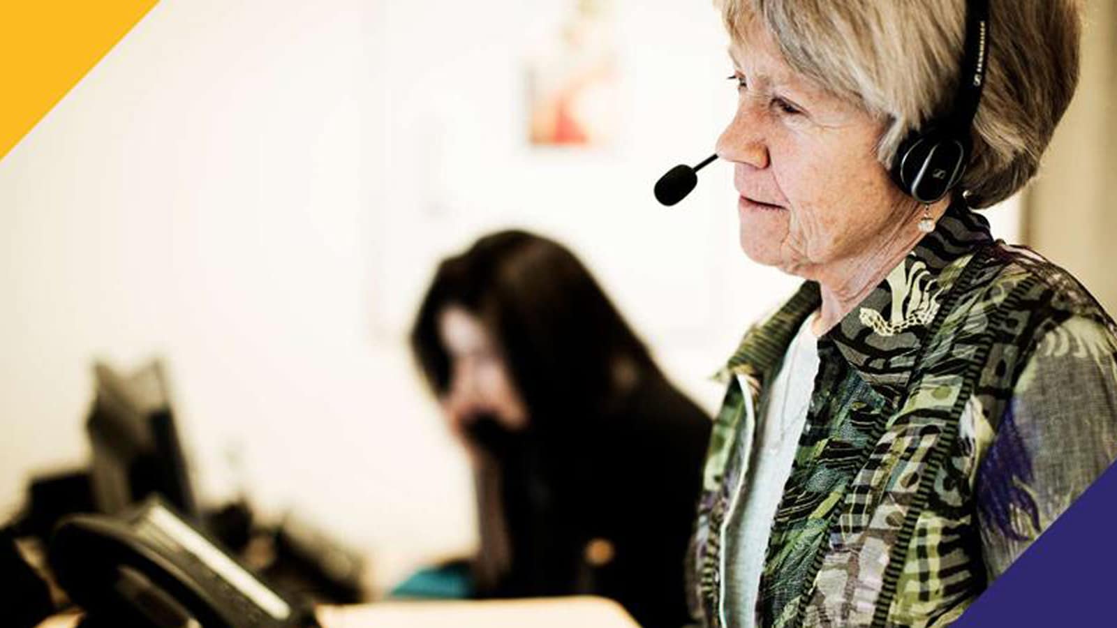 Ældre kvinde med headset på sidder ved et bord, med en mørkhåret kvinde i baggrunden.