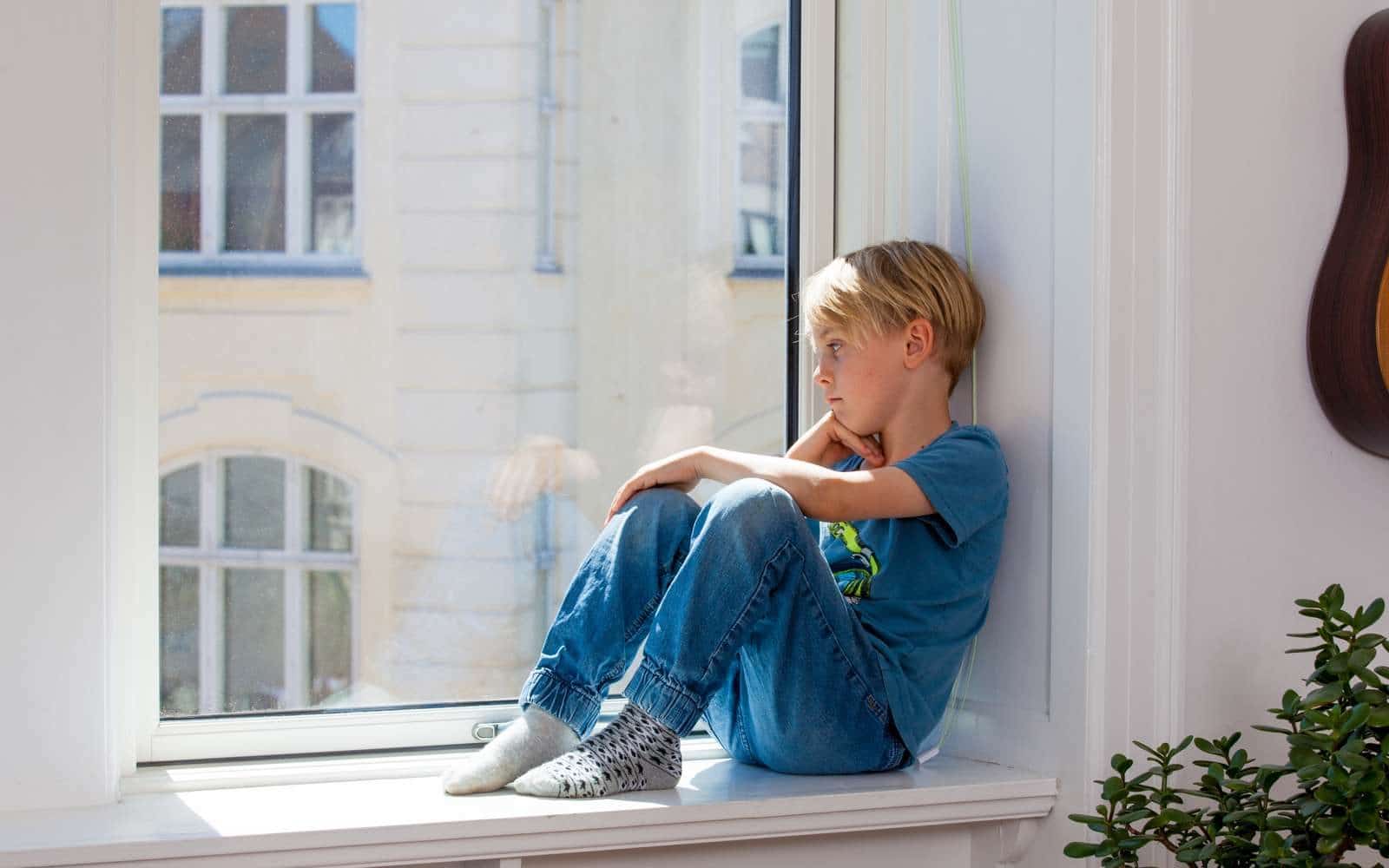 En lyshåret dreng sidder i en vindueskarm og kigger ud på gaden