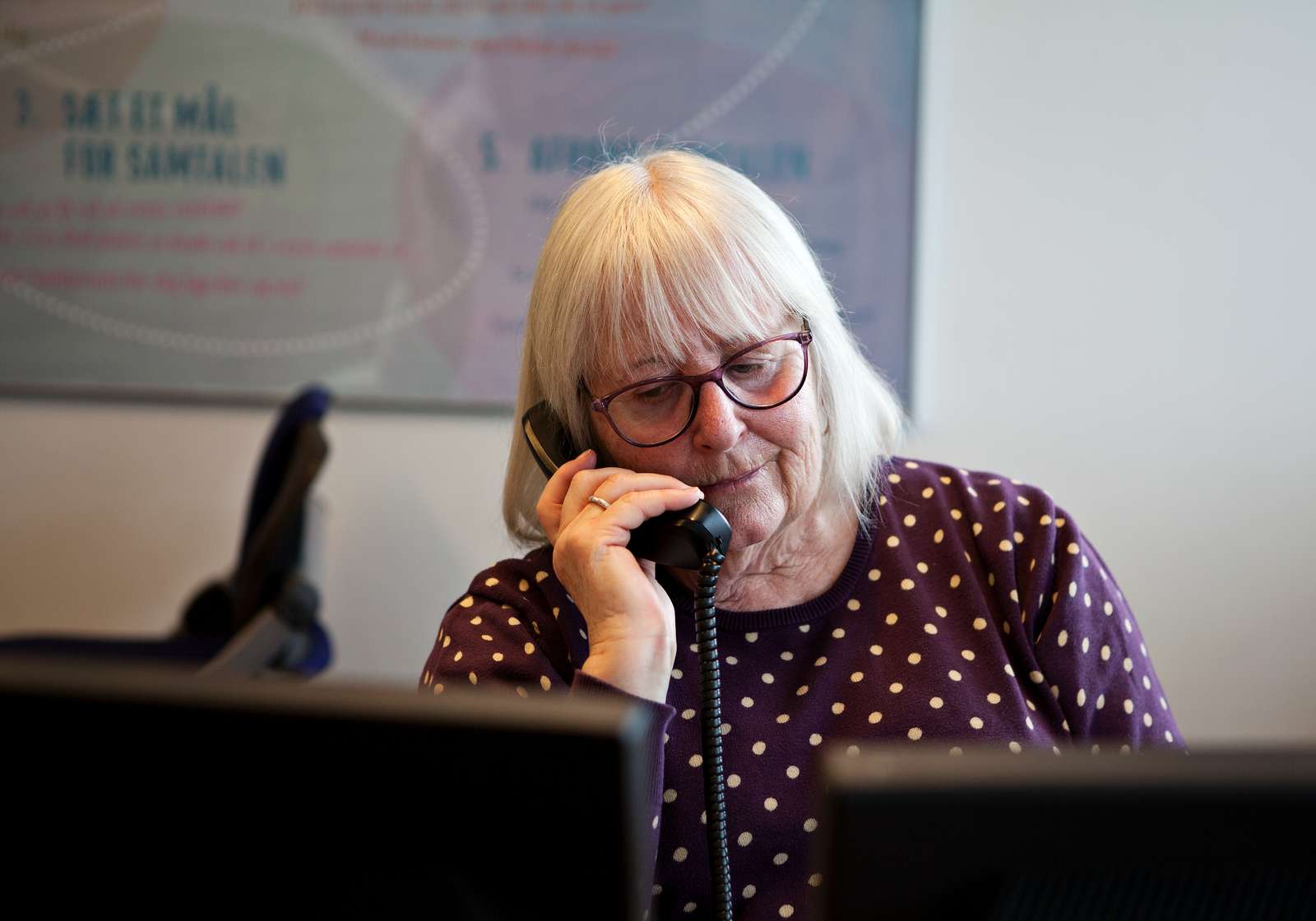 Ældre kvinde med briller taler i telefon foran to skærme.