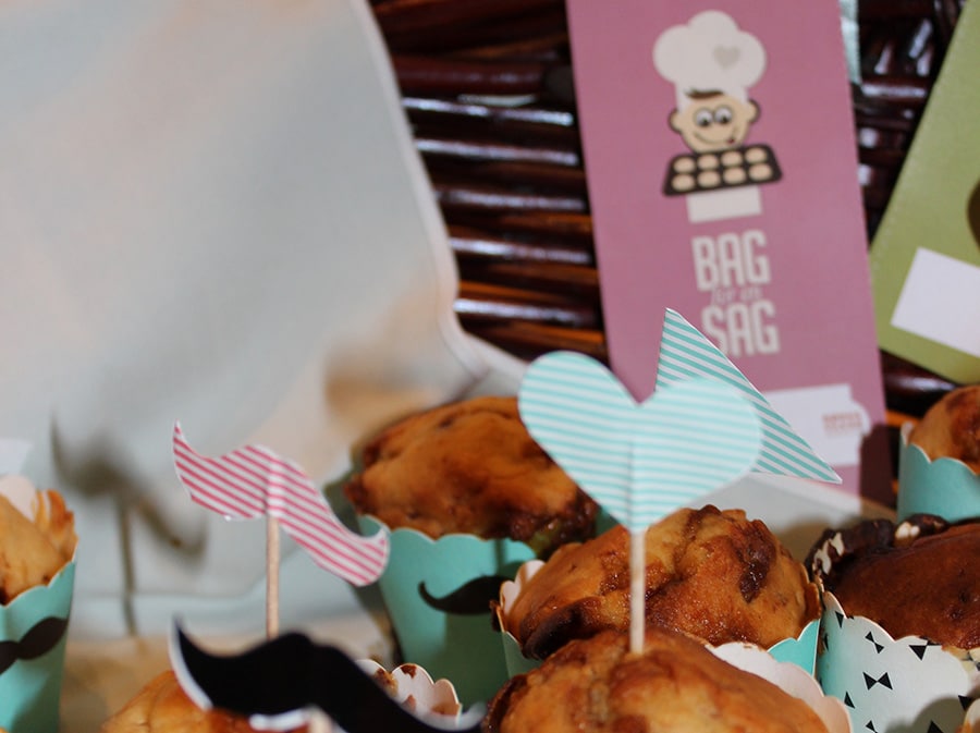 Muffins i forgrunden, med en folder, hvor der står ' BAG FOR EN SAG' på i baggrunden