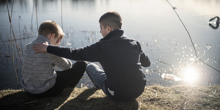 To drenge sidder ved kanten af en sø, hvor den ene dreng trøster den anden