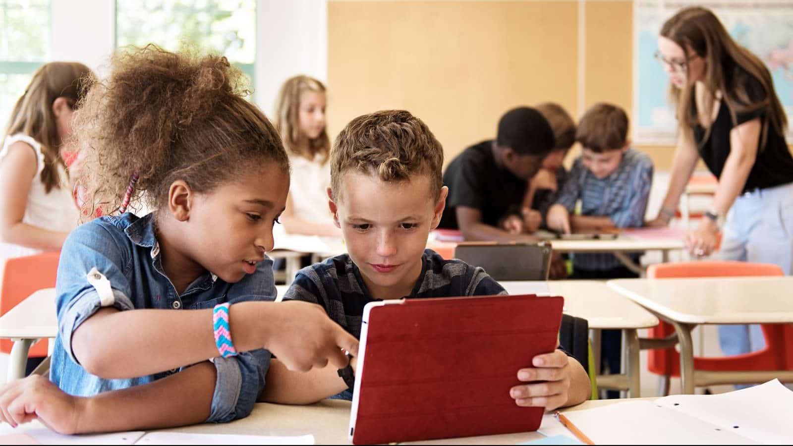 To børn sidder i et klasseværelse og kigger på en iPad.