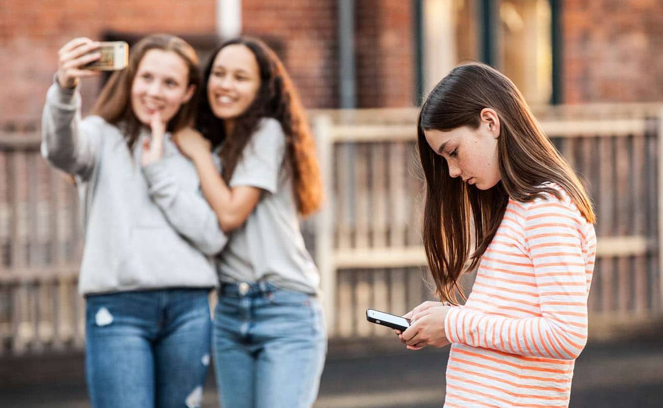 Brunhåret piger kigger ned på sin telefon, mens to piger i baggrunden taget er selfie.