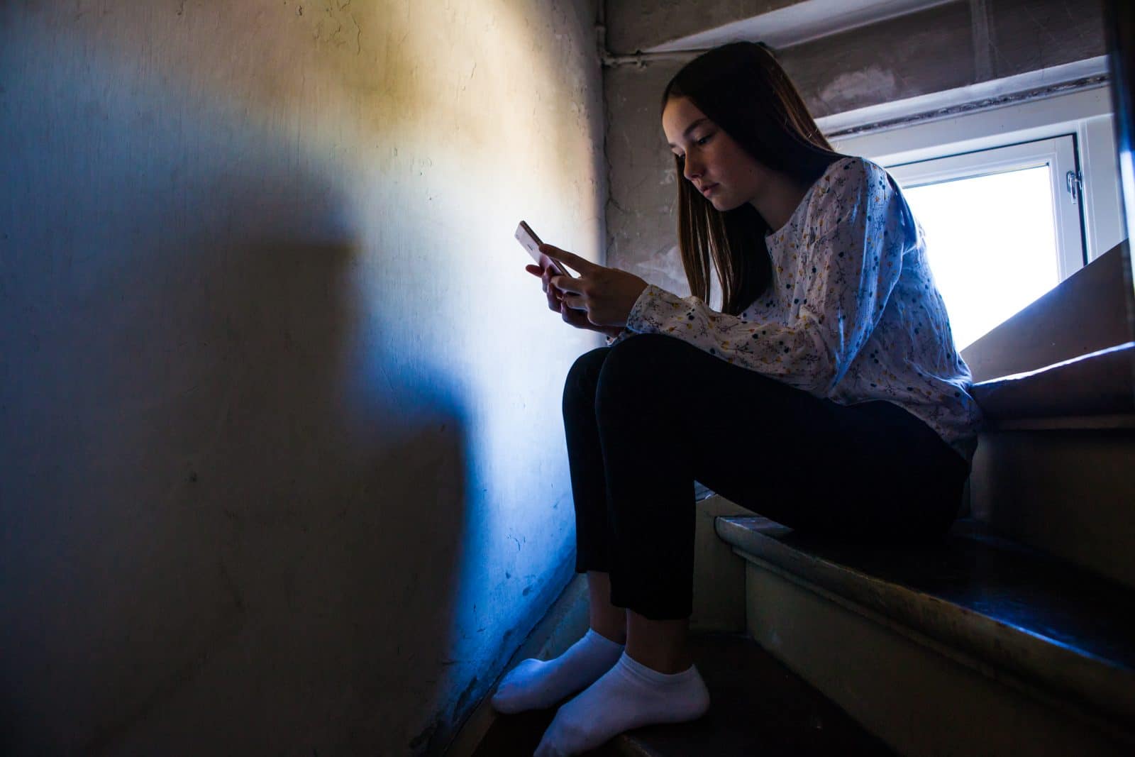 Pige sidder på trappe i mørke med sin mobiltelefon i hånden