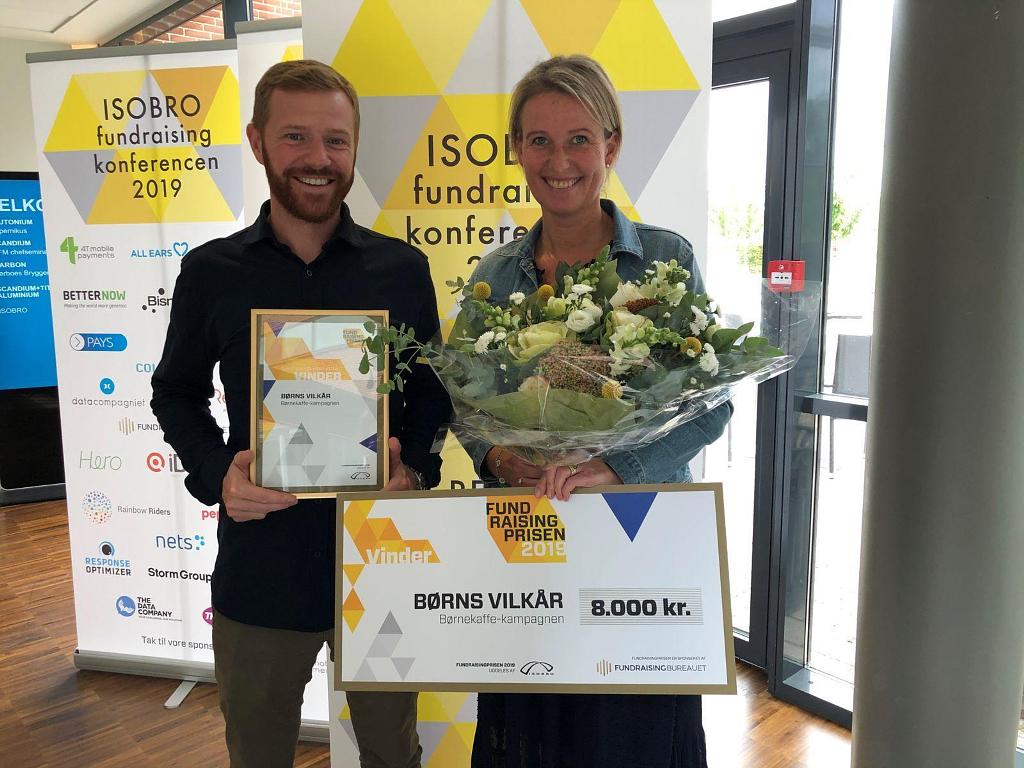 To voksne står og holder fundraisingprisen og en buket blomster