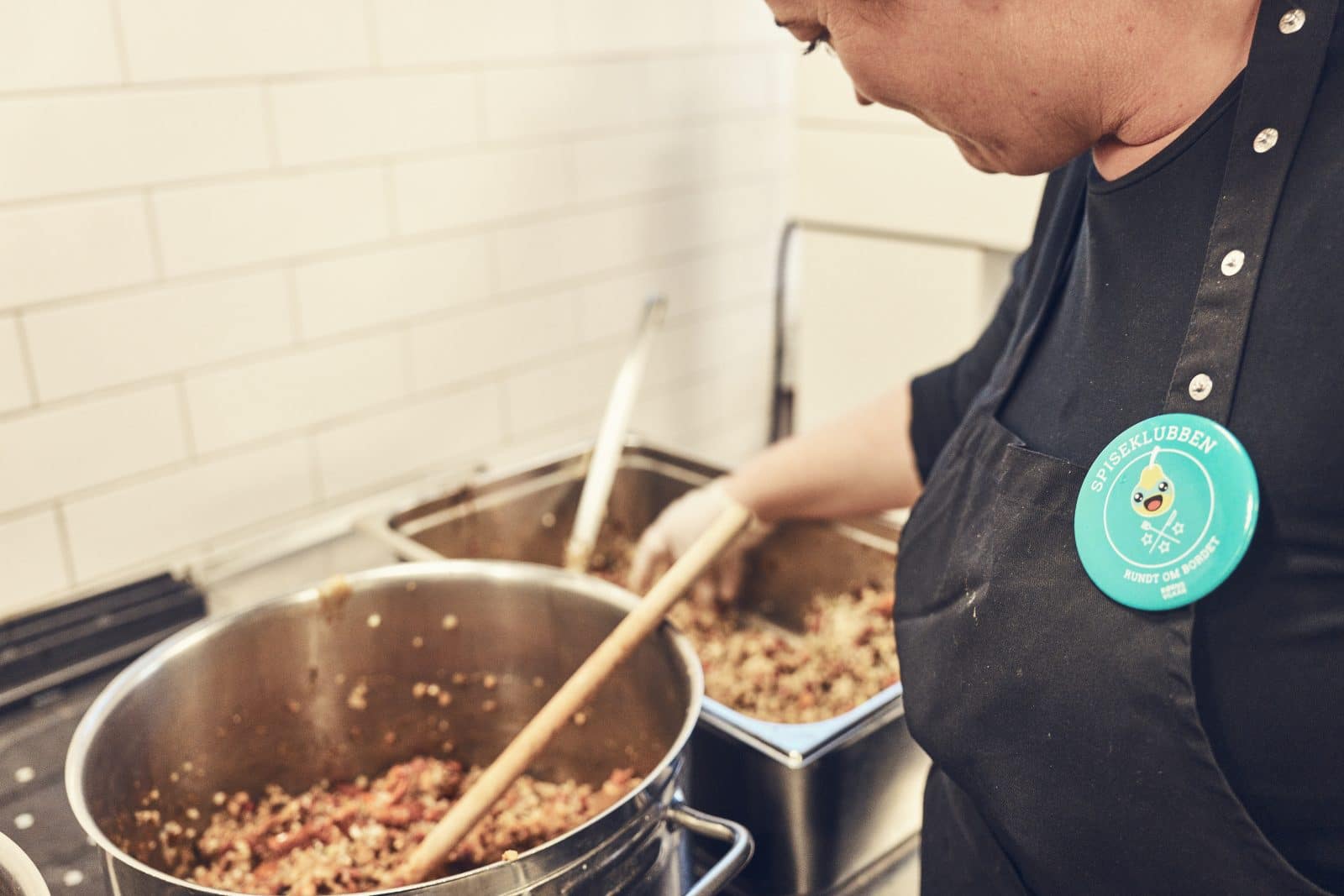 Voksen med et badge for Spiseklubben står over et køkken og laver mad