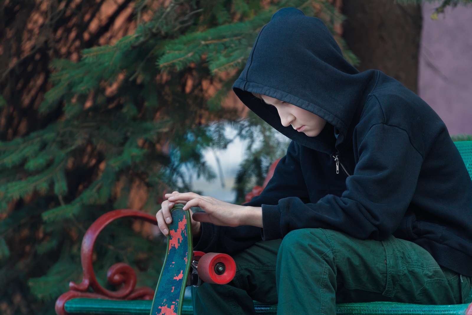 Dreng i hættetrøje med hætten trukket op sidder med et skatebord