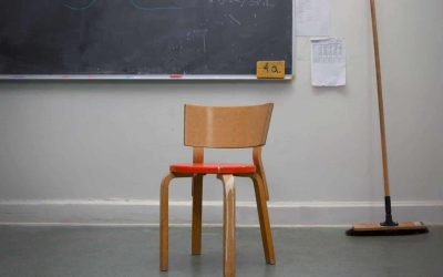 En stol af tre med rødt sæde står i hjørnet af et klasselokale, med en kost og en tavle i baggrunden.