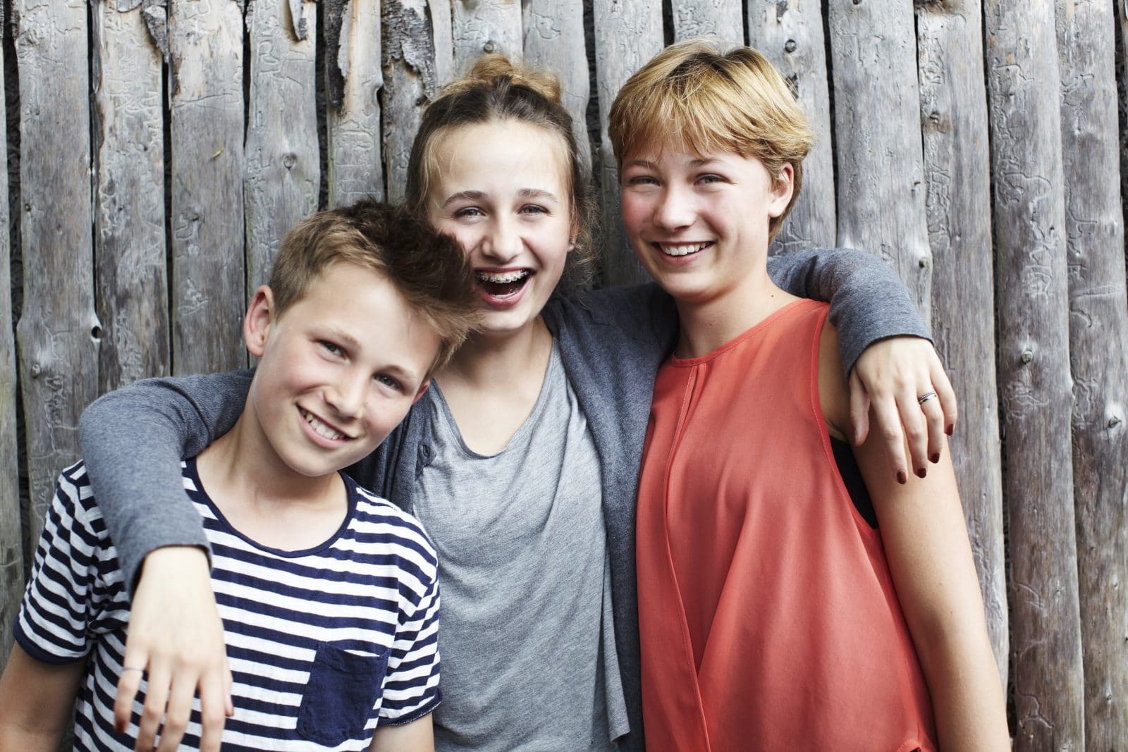 To drenge og en pige griner til kameraet foran et hegn