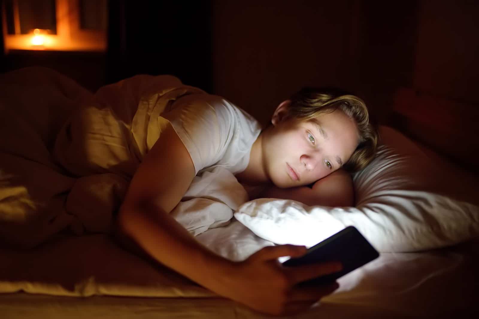 Dreng ligger i sin seng og kigger på sin telefon