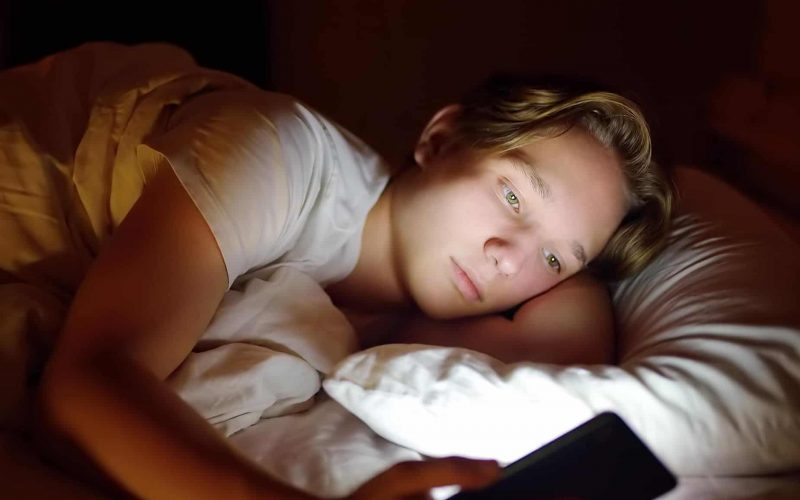 Dreng ligger i sin seng og kigger på sin telefon