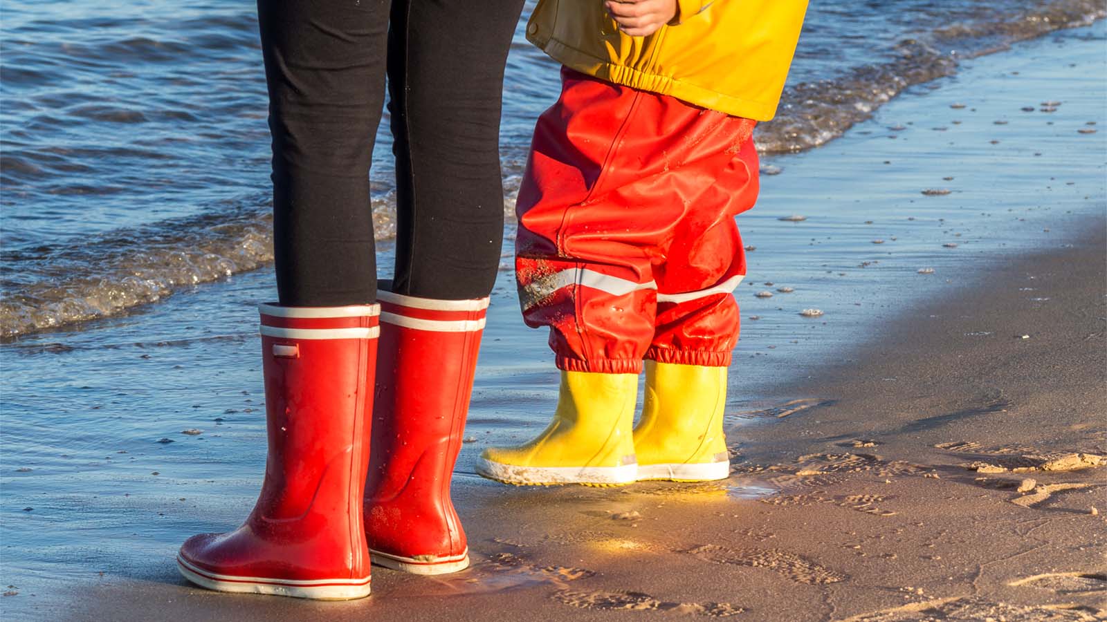 Barn og voksen står på en strand i gummistøvler