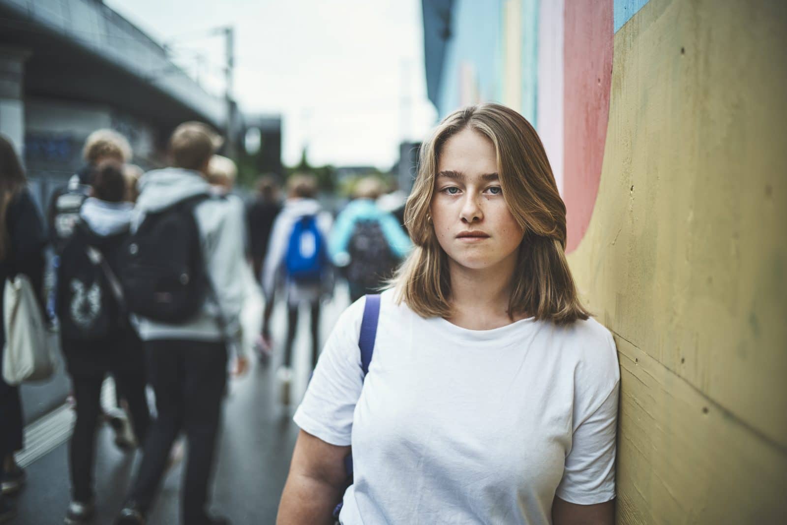 Brunhåret pige står op ad en væg og kigger ind i kameraet.