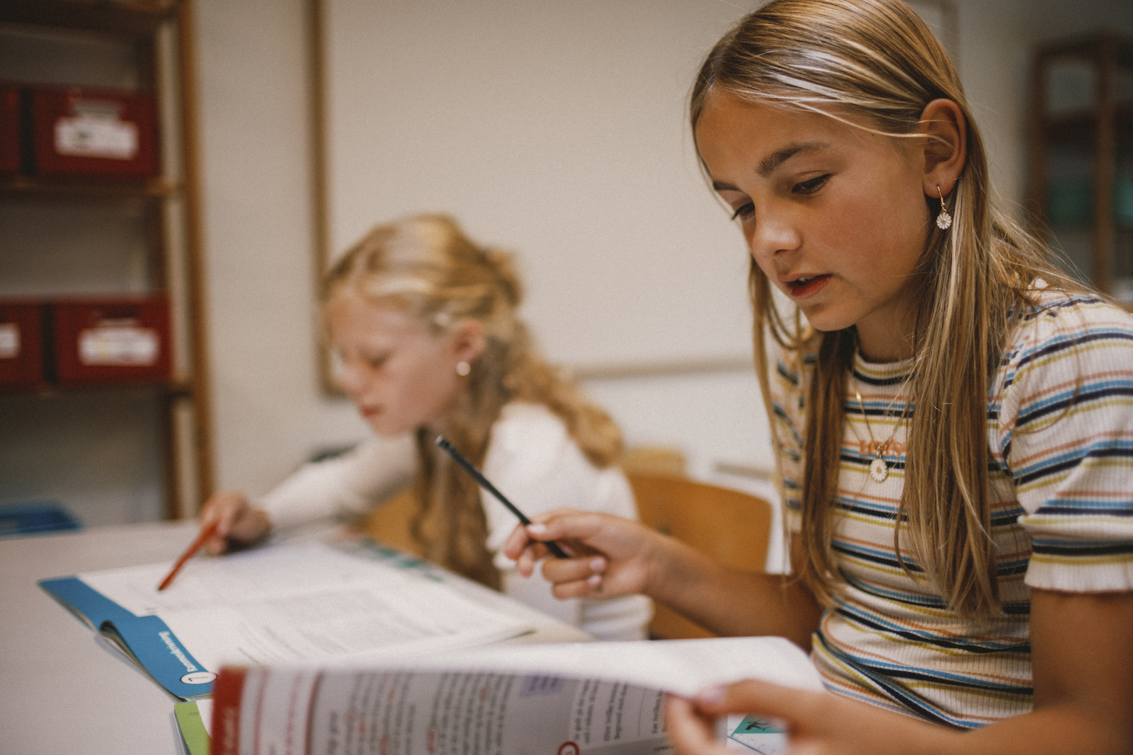 Lyshåret pige læser en skolebog med en anden lyshåret pige i baggrunden
