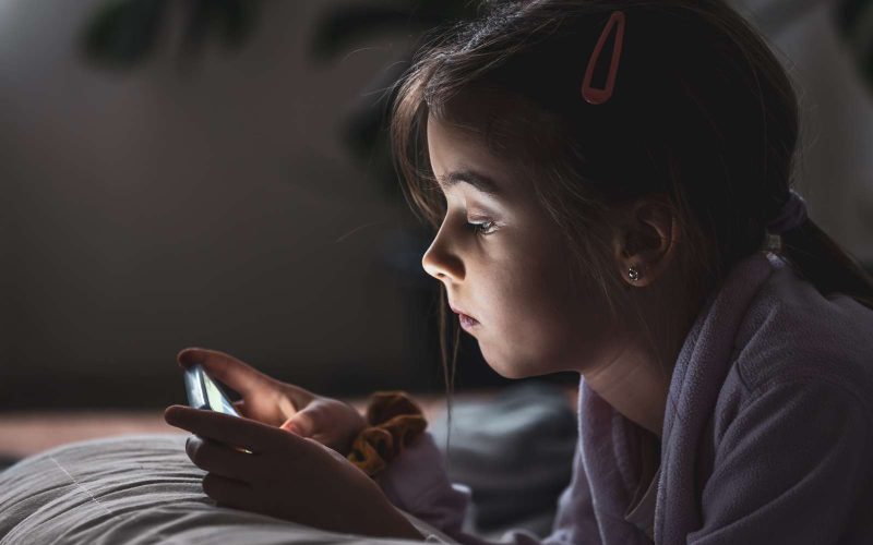Brunhåret pige ligger på en seng, og kigger på en telefon.