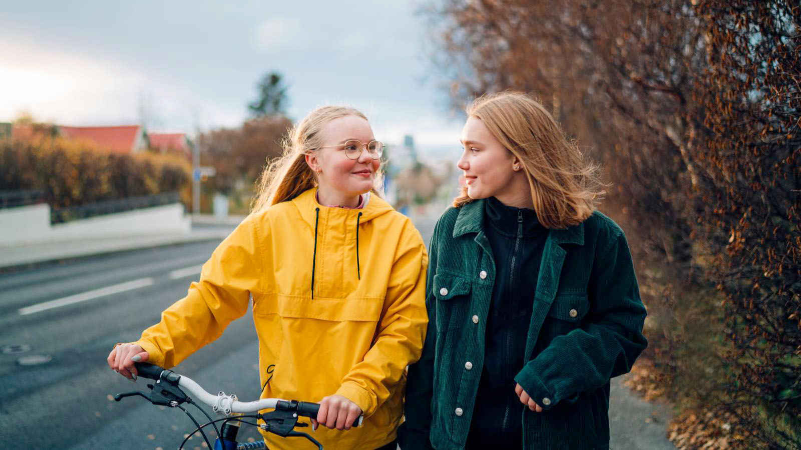 To pige går på en vej og kigger på hinanden, den ene trækker en cykel.