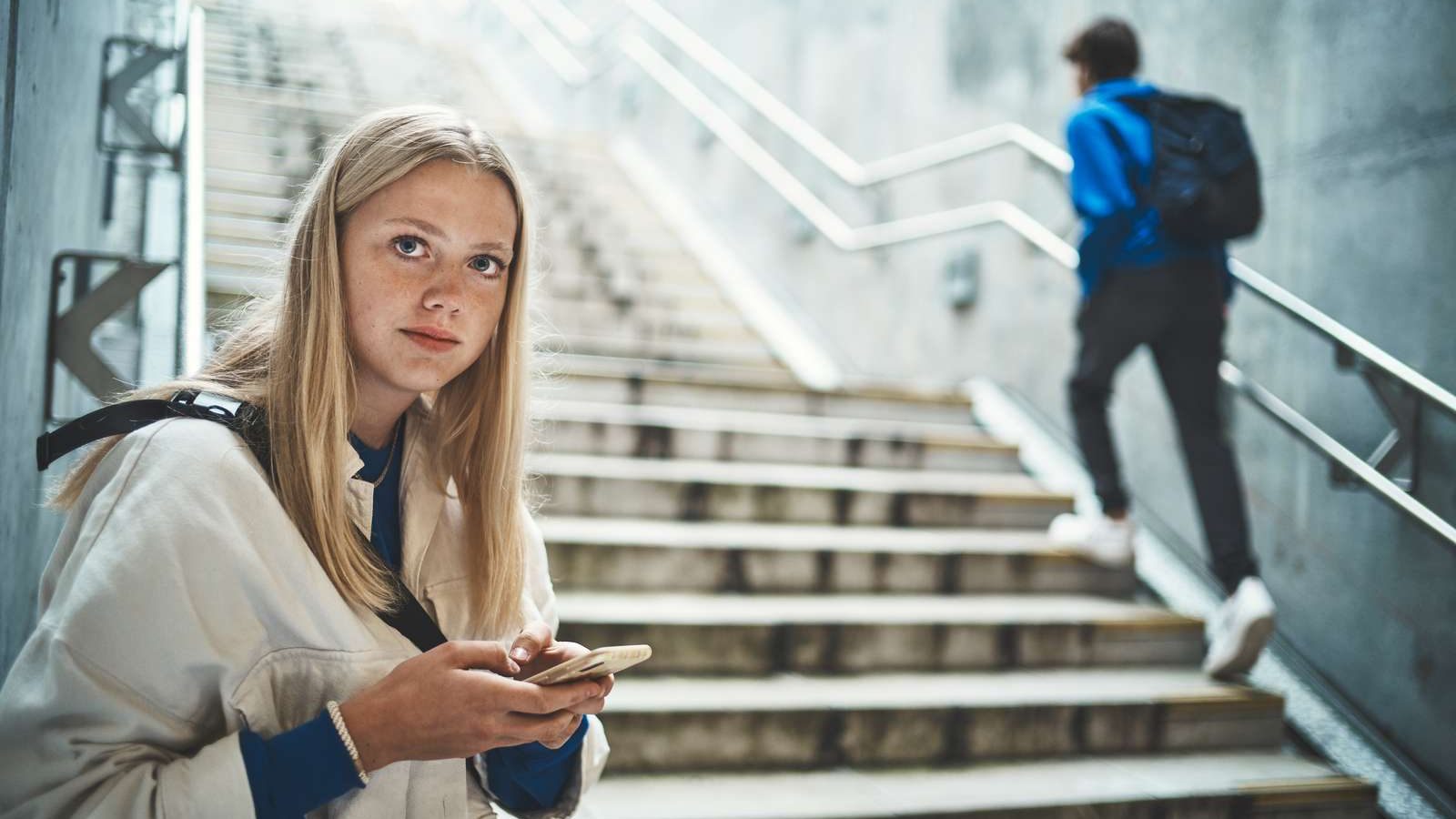 Blond pige for enden af en trappe, med sin telefon i hånden, kigger ind i kameraet