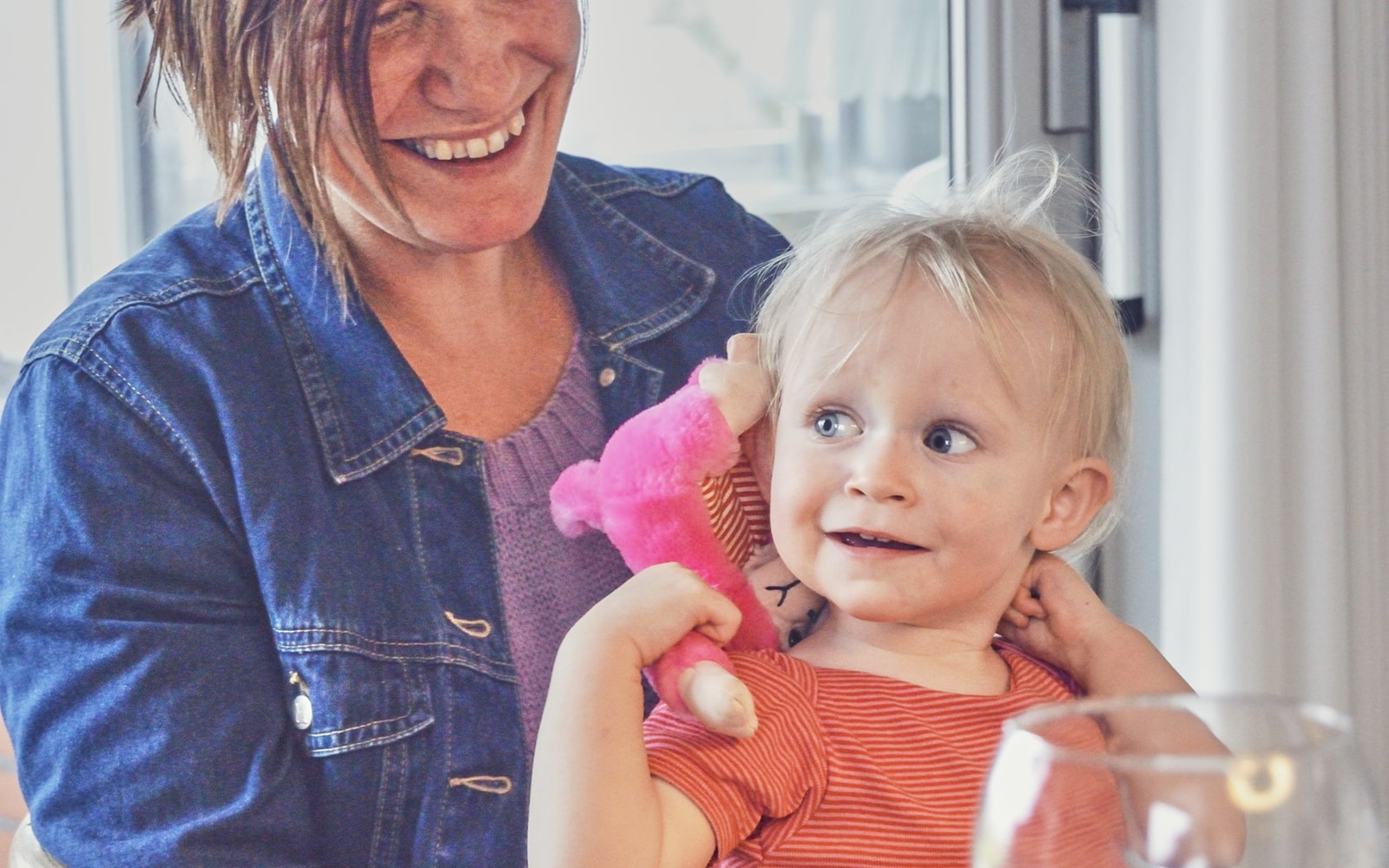 Lyshåret barn holder på en pink bamse, med en brunhåret kvinde i baggrunden