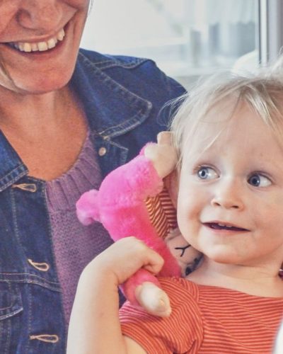 Lyshåret barn holder på en pink bamse, med en brunhåret kvinde i baggrunden
