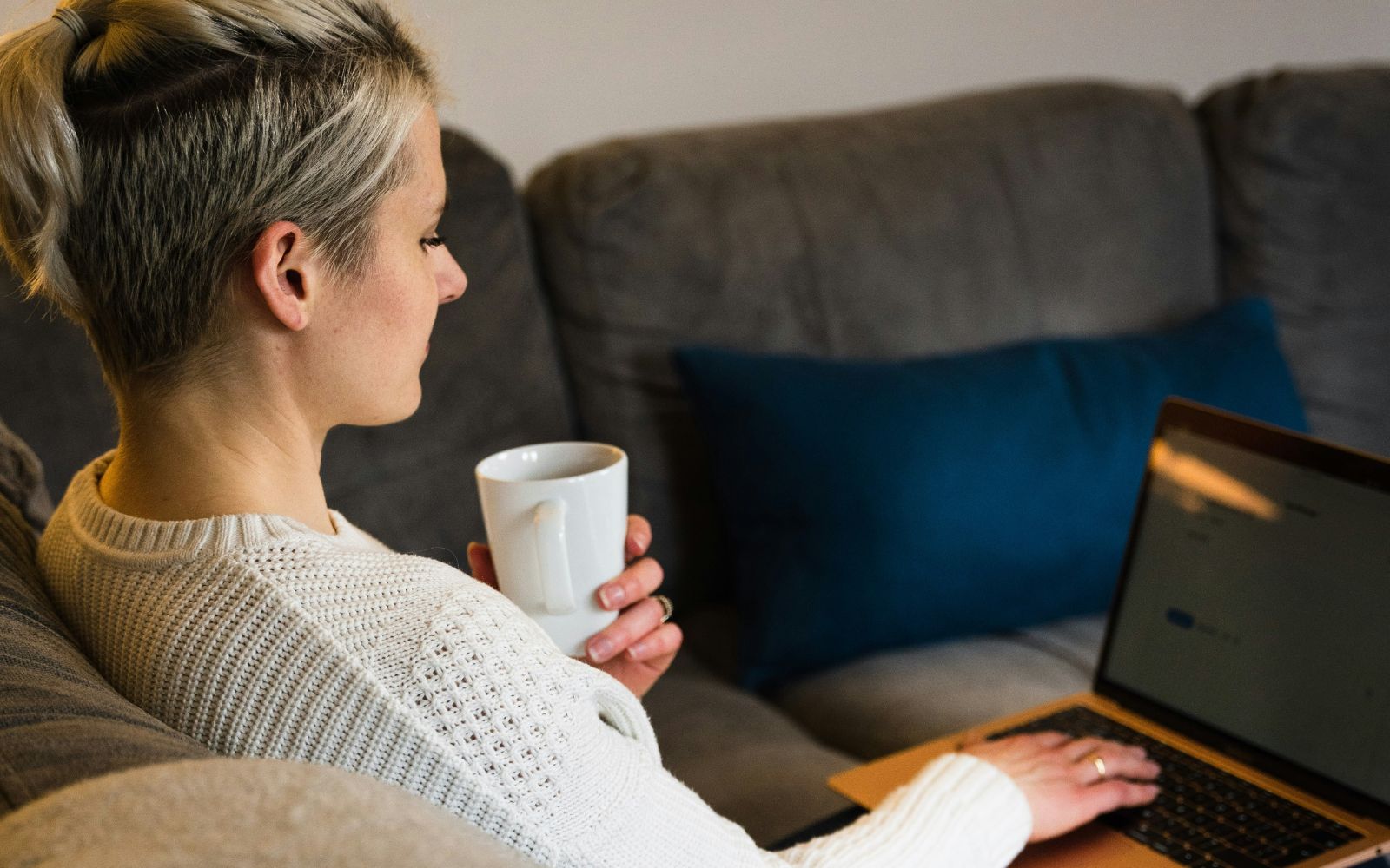 Kvinde med en kop i hånden sidder i sofa med laptop på skødet