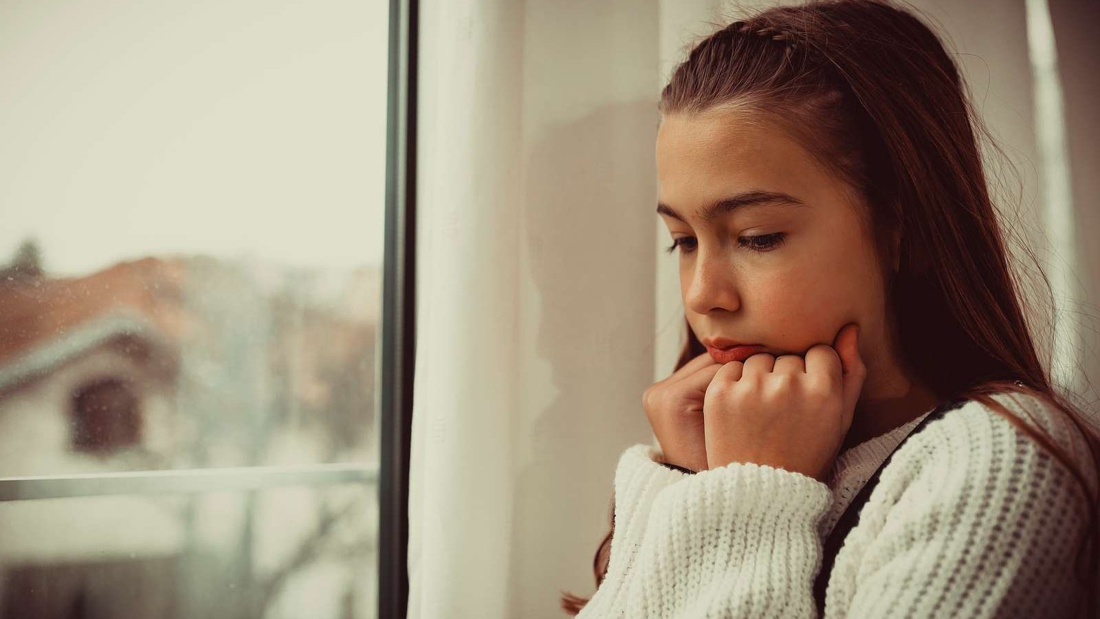 Pige sidder med hagen på hænderne ved siden af et vindue