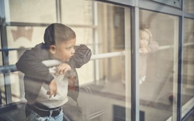 Mørkhåret dreng kigger ud af et vindue, med en lyshåret pige i baggrunden.